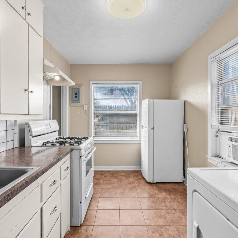 Lubbock Lease Homes: A small kitchen with white cabinets, a gas stove, double sink, white refrigerator, window with blinds, beige walls, tiled floor, and a washing machine next to a window air conditioner.