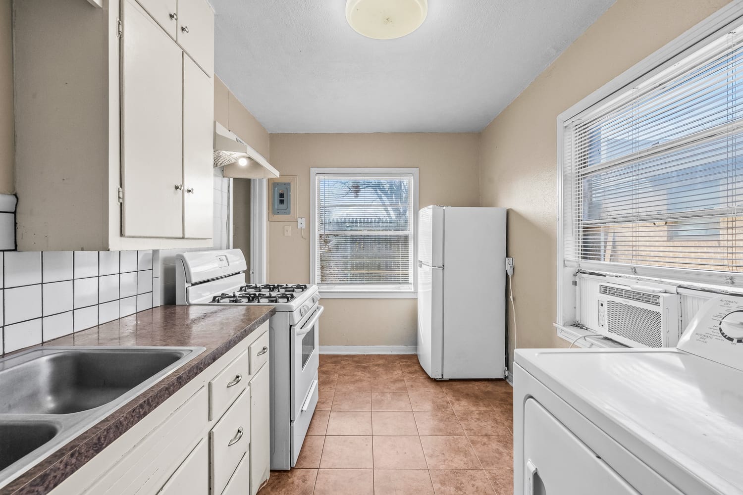 Lubbock Lease Homes: A small kitchen with white cabinets, a gas stove, double sink, white refrigerator, window with blinds, beige walls, tiled floor, and a washing machine next to a window air conditioner.