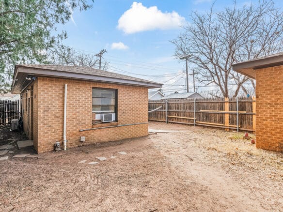 Lubbock Lease Homes: A backyard with a brick house featuring a window air conditioner, dry patchy ground, a wooden fence, leafless trees, and a clear blue sky with a few clouds.