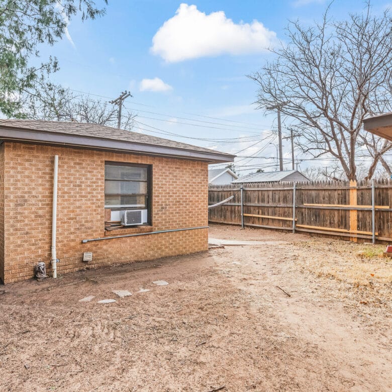Lubbock Lease Homes: A backyard with a brick house featuring a window air conditioner, dry patchy ground, a wooden fence, leafless trees, and a clear blue sky with a few clouds.