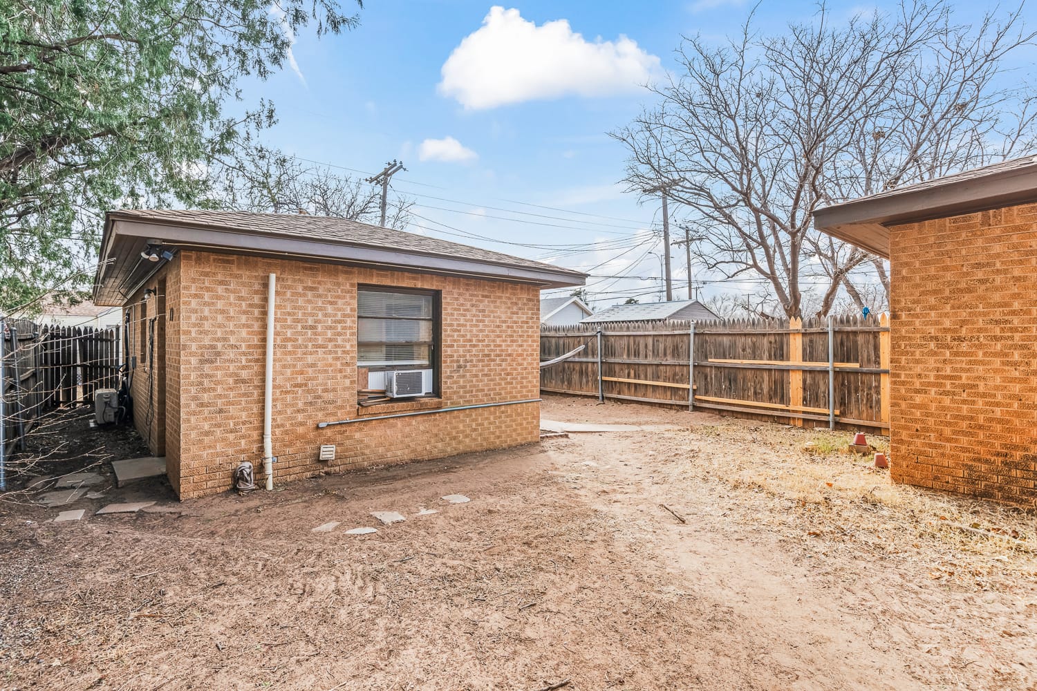 Lubbock Lease Homes: A backyard with a brick house featuring a window air conditioner, dry patchy ground, a wooden fence, leafless trees, and a clear blue sky with a few clouds.