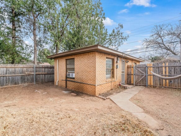 Lubbock Lease Homes: A small brick house with an air conditioner in the window sits in a fenced backyard with dry grass, trees, a hammock, and a stone path leading to a wooden gate under a blue sky.