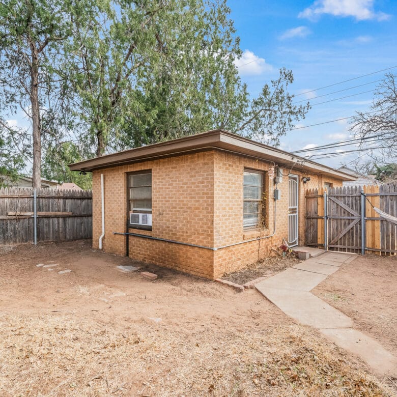 Lubbock Lease Homes: A small brick house with an air conditioner in the window sits in a fenced backyard with dry grass, trees, a hammock, and a stone path leading to a wooden gate under a blue sky.