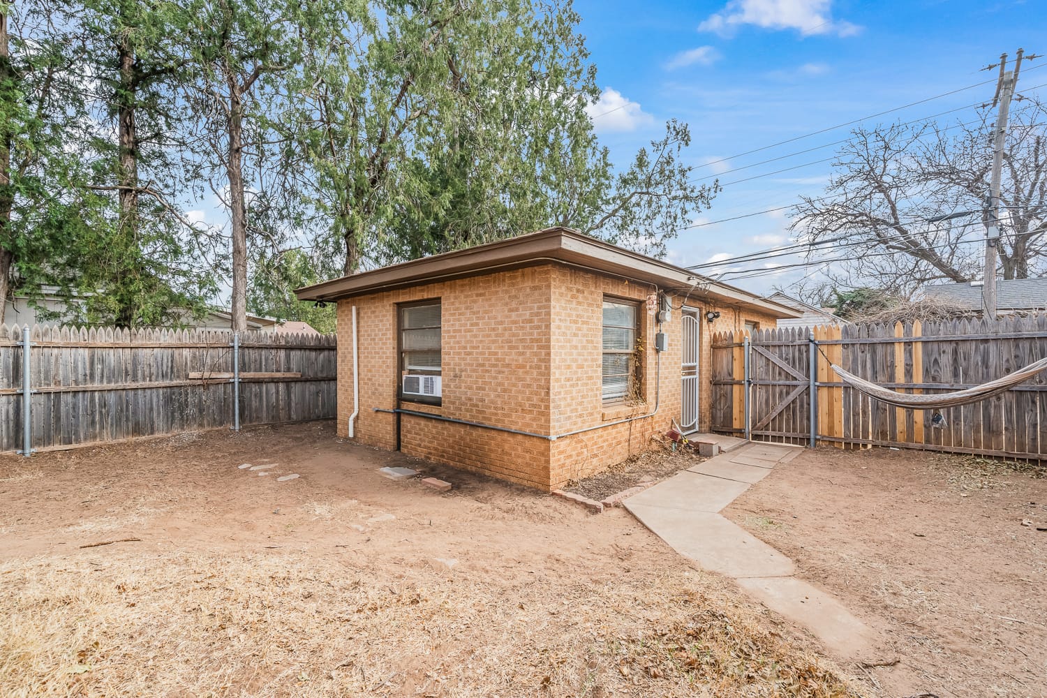 Lubbock Lease Homes: A small brick house with an air conditioner in the window sits in a fenced backyard with dry grass, trees, a hammock, and a stone path leading to a wooden gate under a blue sky.