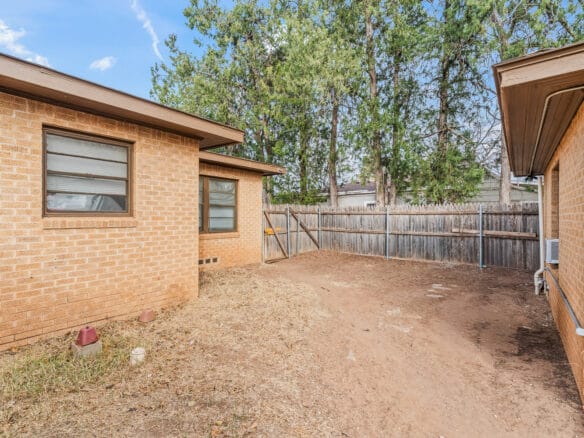 Lubbock Lease Homes: A backyard with a dirt ground, brick house walls on two sides, a wooden fence, some small plants, and tall trees in the background under a partly cloudy sky.