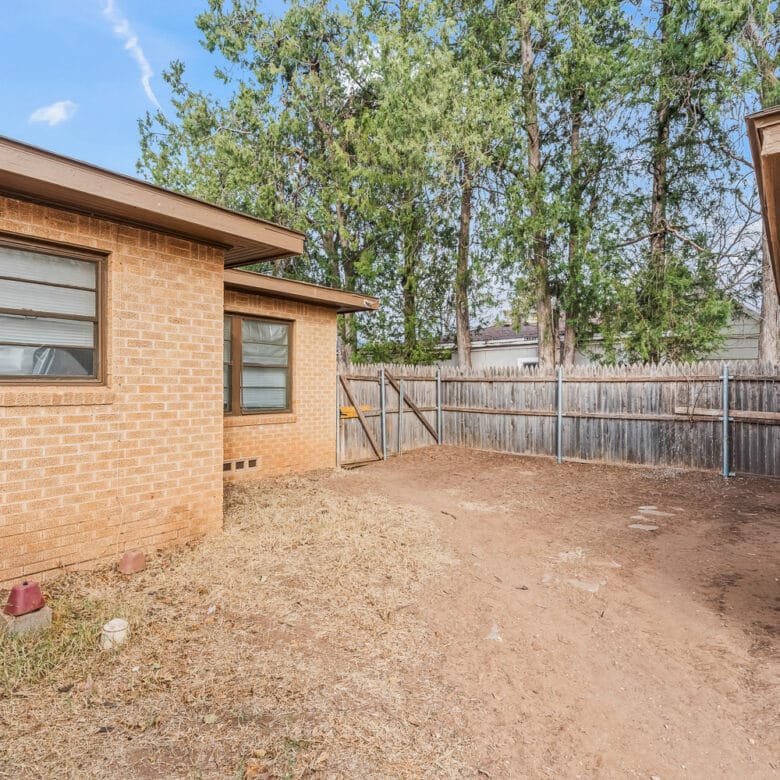 Lubbock Lease Homes: A backyard with a dirt ground, brick house walls on two sides, a wooden fence, some small plants, and tall trees in the background under a partly cloudy sky.