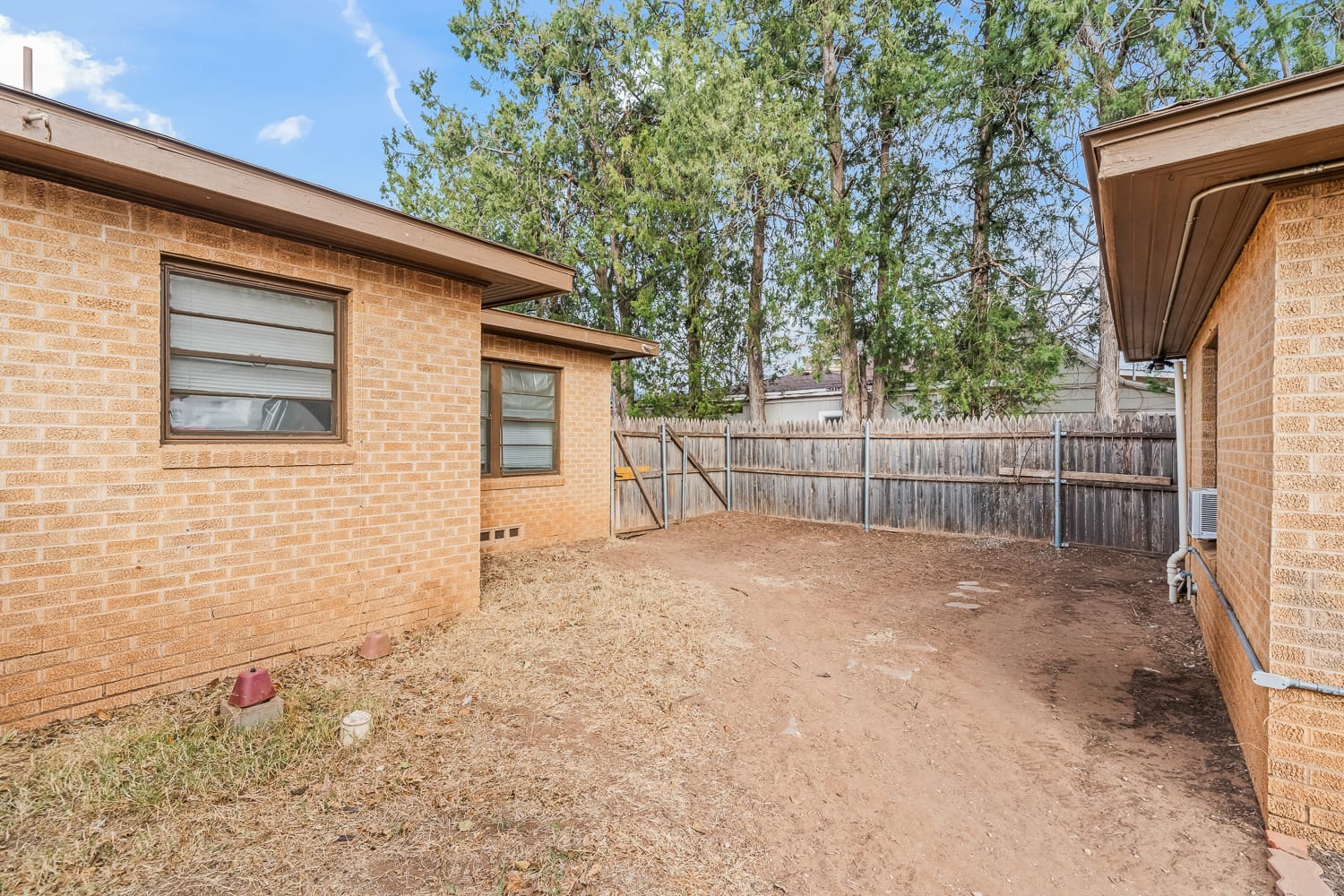 Lubbock Lease Homes: A backyard with a dirt ground, brick house walls on two sides, a wooden fence, some small plants, and tall trees in the background under a partly cloudy sky.