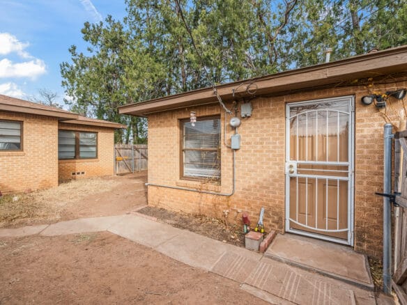 Lubbock Lease Homes: A tan brick house with a metal screen door and large window, surrounded by dirt and a concrete walkway, with another similar house and tall trees in the background.