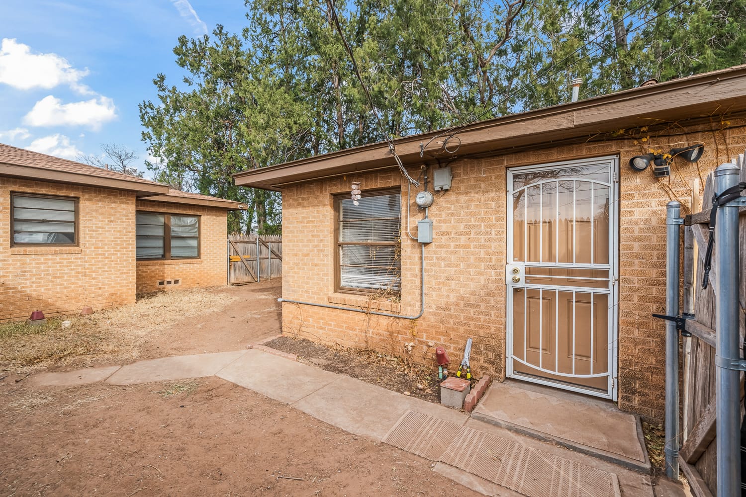 Lubbock Lease Homes: A tan brick house with a metal screen door and large window, surrounded by dirt and a concrete walkway, with another similar house and tall trees in the background.