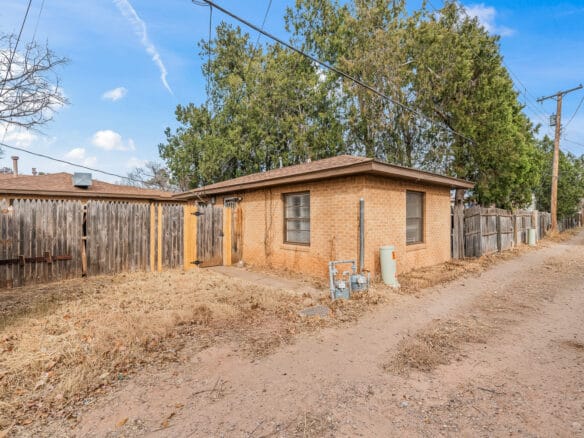 Lubbock Lease Homes: A small, single-story brick house with a fenced backyard sits beside a dirt alleyway. Tall trees and wooden fences surround the property under a blue sky with scattered clouds.