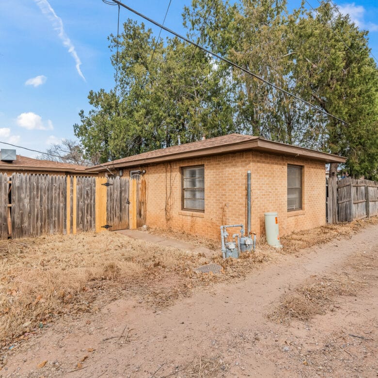 Lubbock Lease Homes: A small, single-story brick house with a fenced backyard sits beside a dirt alleyway. Tall trees and wooden fences surround the property under a blue sky with scattered clouds.