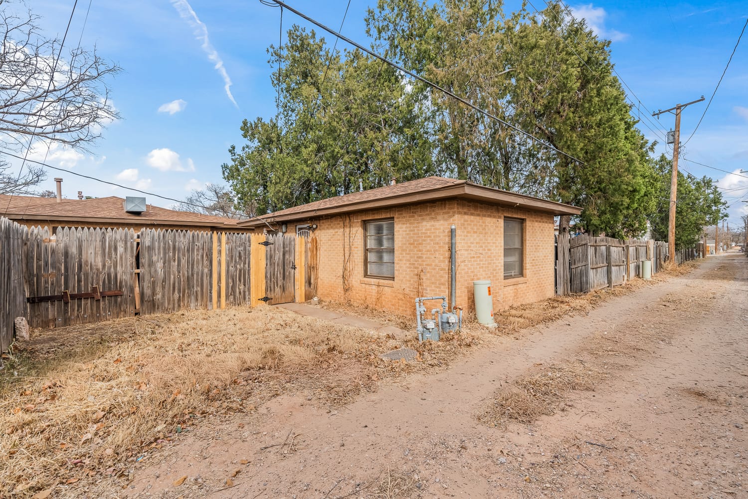 Lubbock Lease Homes: A small, single-story brick house with a fenced backyard sits beside a dirt alleyway. Tall trees and wooden fences surround the property under a blue sky with scattered clouds.