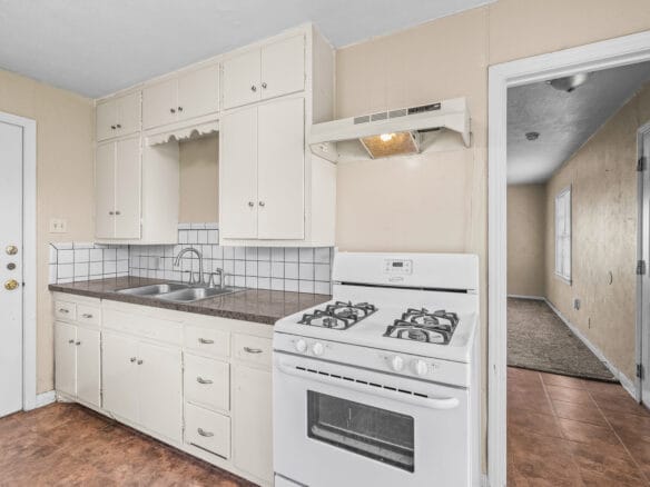 Lubbock Lease Homes: Bright kitchen with white cabinets, gas stove, sink, and brown countertop. White tile backsplash, beige walls, and brown tile floor. Open doorway leads to a carpeted hallway with natural light.