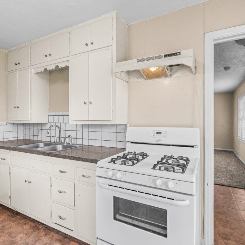 Lubbock Lease Homes: Bright kitchen with white cabinets, gas stove, sink, and brown countertop. White tile backsplash, beige walls, and brown tile floor. Open doorway leads to a carpeted hallway with natural light.
