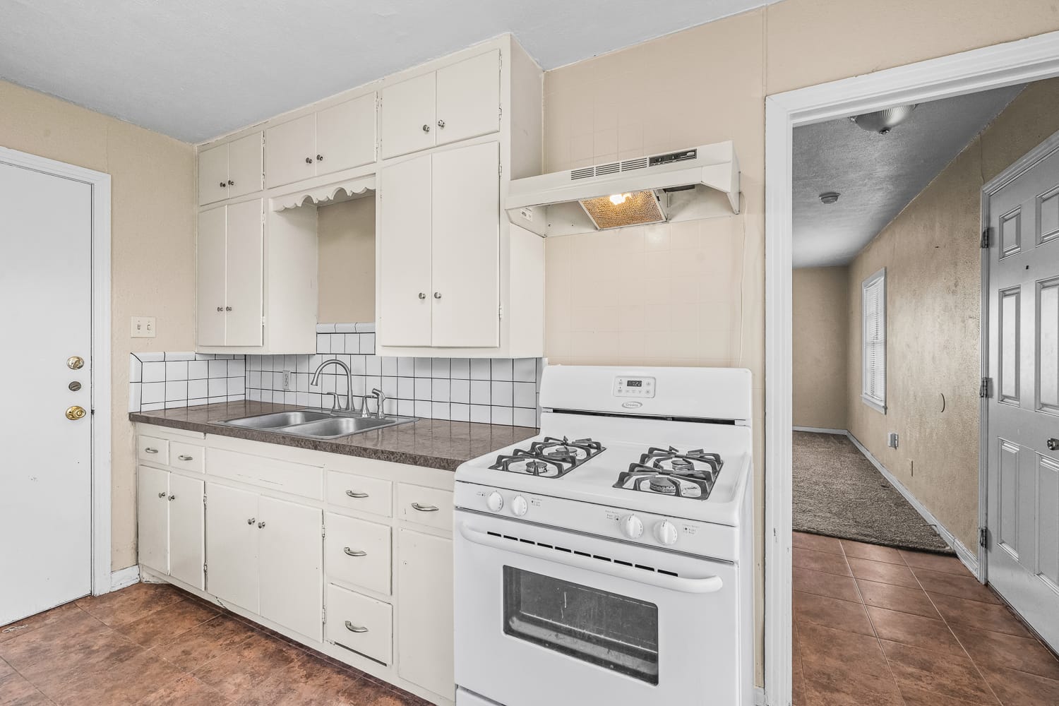 Lubbock Lease Homes: Bright kitchen with white cabinets, gas stove, sink, and brown countertop. White tile backsplash, beige walls, and brown tile floor. Open doorway leads to a carpeted hallway with natural light.