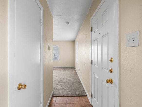 Lubbock Lease Homes: A view of a narrow entry hallway with a white front door, beige walls, a closet door on the left, tiled flooring near the entrance, and carpeted flooring leading to a room with two windows.