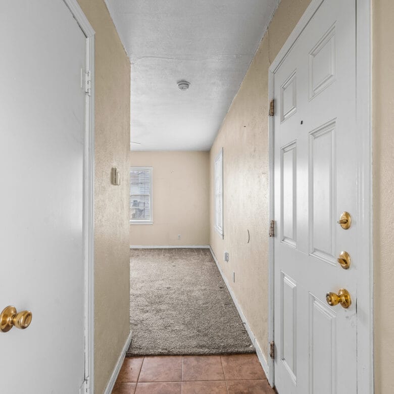 Lubbock Lease Homes: A view of a narrow entry hallway with a white front door, beige walls, a closet door on the left, tiled flooring near the entrance, and carpeted flooring leading to a room with two windows.
