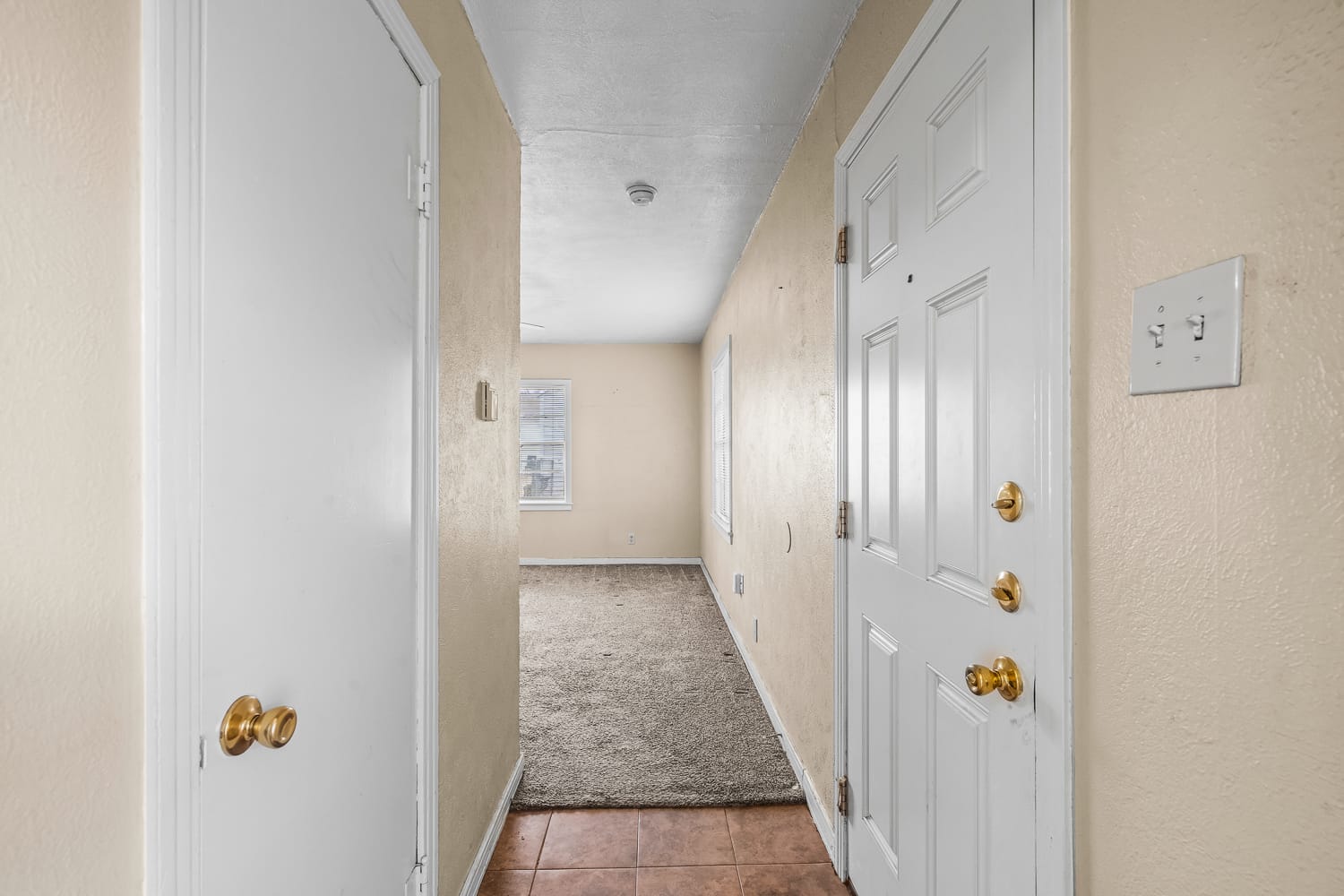Lubbock Lease Homes: A view of a narrow entry hallway with a white front door, beige walls, a closet door on the left, tiled flooring near the entrance, and carpeted flooring leading to a room with two windows.