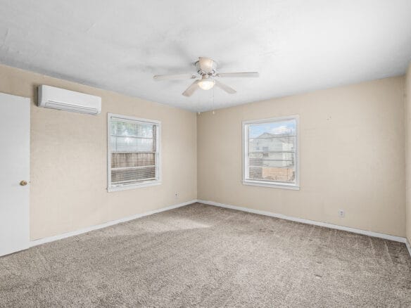 Lubbock Lease Homes: A spacious, empty room with beige walls, three windows, carpeted floor, ceiling fan with a light, and a wall-mounted air conditioning unit. Natural light fills the room through the windows.