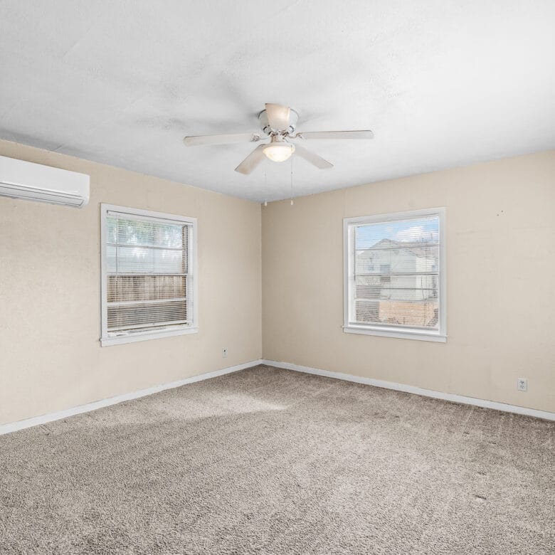 Lubbock Lease Homes: A spacious, empty room with beige walls, three windows, carpeted floor, ceiling fan with a light, and a wall-mounted air conditioning unit. Natural light fills the room through the windows.