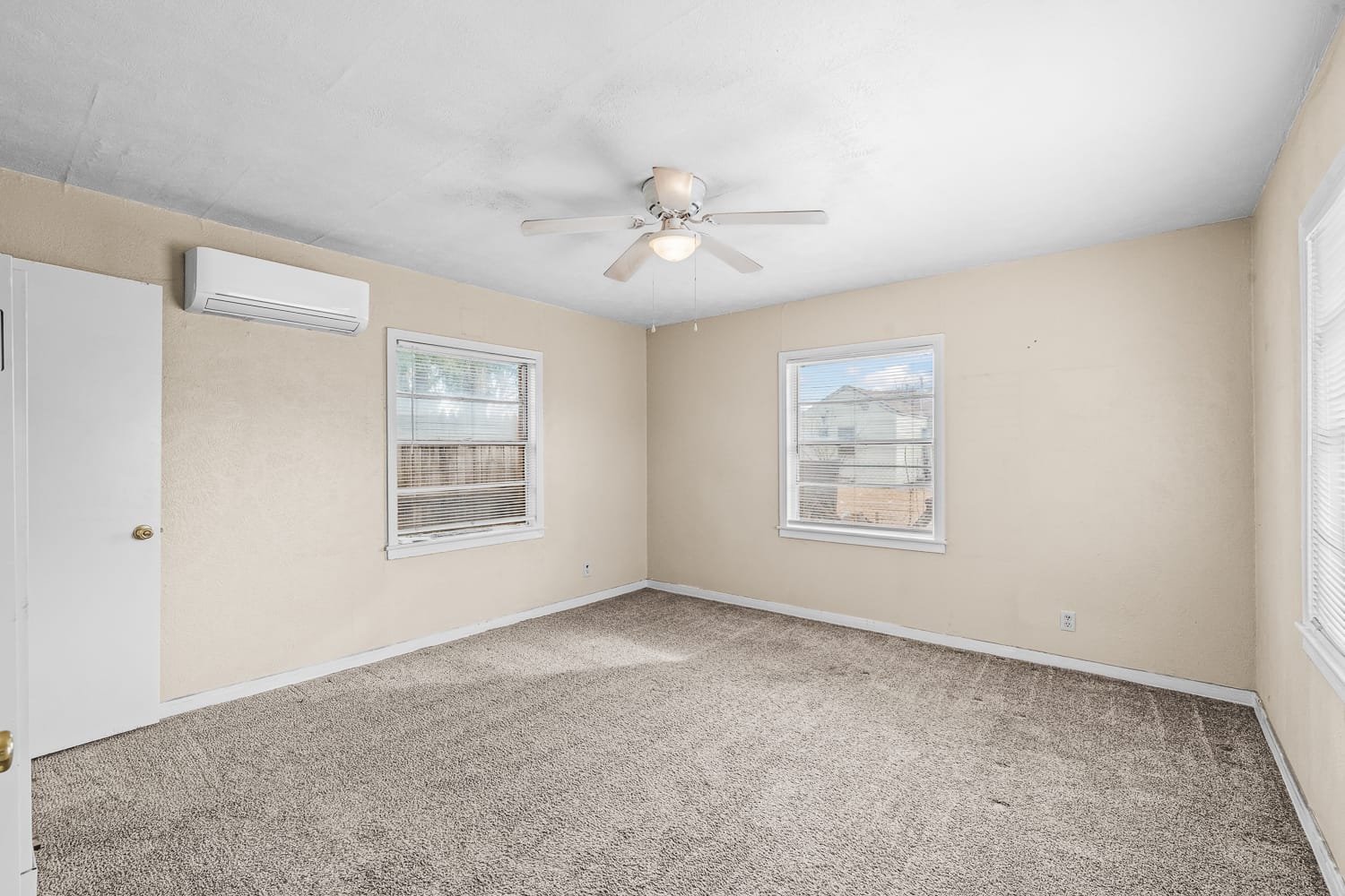 Lubbock Lease Homes: A spacious, empty room with beige walls, three windows, carpeted floor, ceiling fan with a light, and a wall-mounted air conditioning unit. Natural light fills the room through the windows.