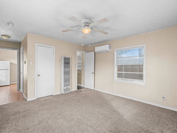 Lubbock Lease Homes: A beige carpeted living room with a ceiling fan, two windows, and several doors; a small hallway leads to a kitchen area with a white refrigerator visible.
