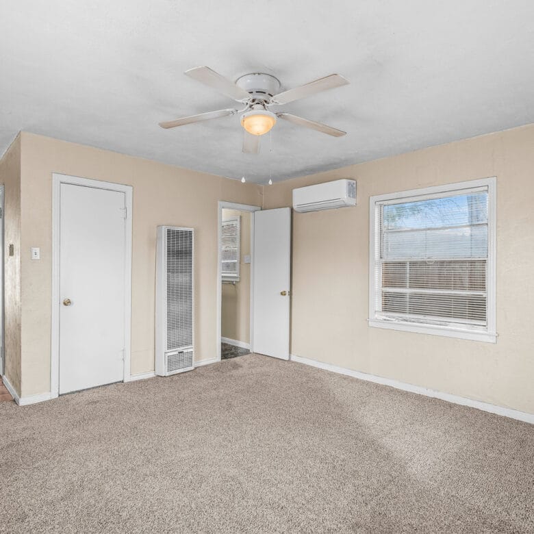 Lubbock Lease Homes: A beige carpeted living room with a ceiling fan, two windows, and several doors; a small hallway leads to a kitchen area with a white refrigerator visible.