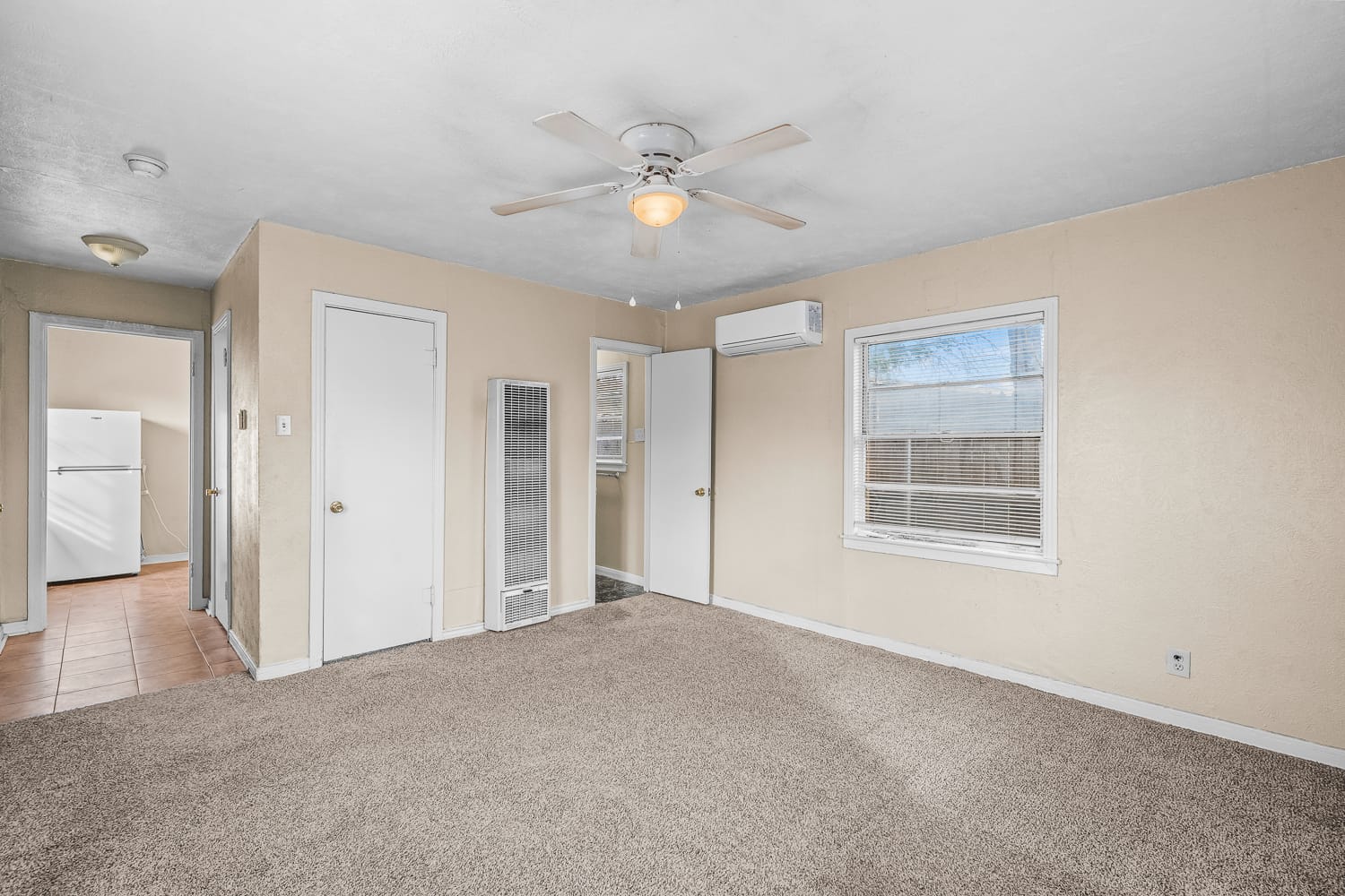 Lubbock Lease Homes: A beige carpeted living room with a ceiling fan, two windows, and several doors; a small hallway leads to a kitchen area with a white refrigerator visible.