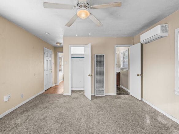 Lubbock Lease Homes: A bright, beige-walled room with carpeted floor, ceiling fan with light, open closet and doors, wall heater, air conditioning unit, and several windows letting in natural light.