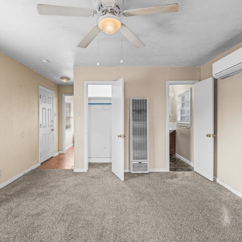 Lubbock Lease Homes: A bright, beige-walled room with carpeted floor, ceiling fan with light, open closet and doors, wall heater, air conditioning unit, and several windows letting in natural light.