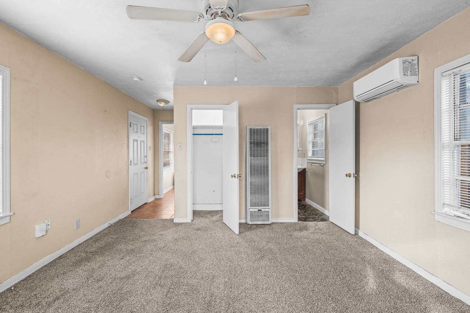 Lubbock Lease Homes: A bright, beige-walled room with carpeted floor, ceiling fan with light, open closet and doors, wall heater, air conditioning unit, and several windows letting in natural light.
