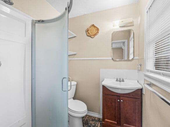 Lubbock Lease Homes: Small bathroom with a frosted glass corner shower, white toilet, wooden vanity with sink, wall-mounted mirror, shelves, and a window with blinds letting in light. The walls are beige and the floor has a dark pattern.