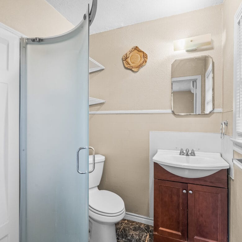 Lubbock Lease Homes: Small bathroom with a frosted glass corner shower, white toilet, wooden vanity with sink, wall-mounted mirror, shelves, and a window with blinds letting in light. The walls are beige and the floor has a dark pattern.
