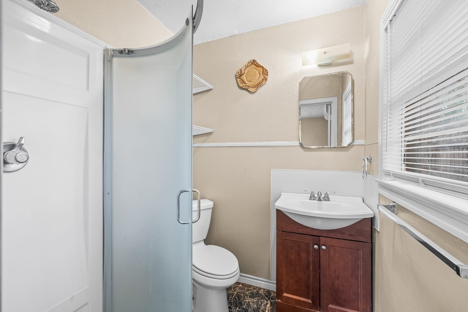 Lubbock Lease Homes: Small bathroom with a frosted glass corner shower, white toilet, wooden vanity with sink, wall-mounted mirror, shelves, and a window with blinds letting in light. The walls are beige and the floor has a dark pattern.