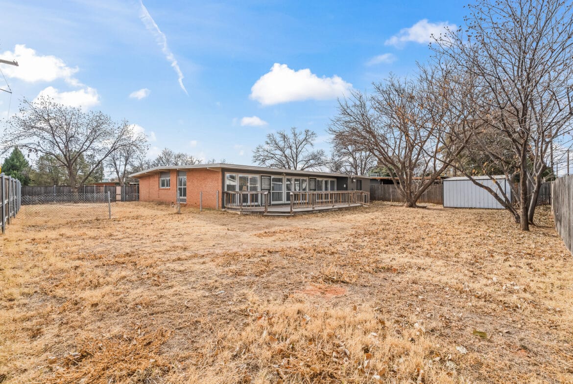 Lubbock Lease Homes: A single-story brick house with a fenced yard, bare trees, a wooden ramp leading to the back entrance, and a storage shed, all under a bright blue sky with a few clouds.